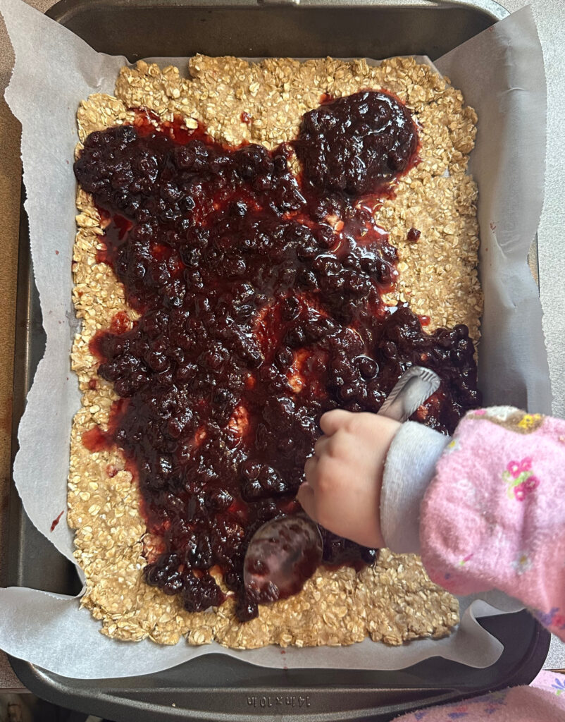 a child's had spreading jam over a sourdough discard and oatmeal crust