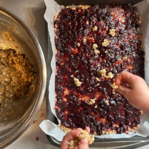 Child sprinkling oatmeal topping onto the jam layer of sourdough discard jam bars.